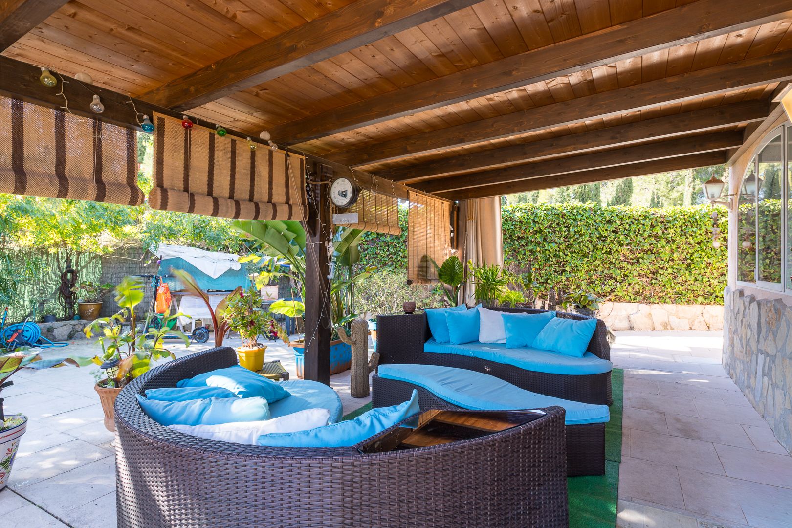 Terraza cubierta con pérgola de madera oscura, equipada con sofás de ratán curvos y cojines azules, rodeada de vegetación y persianas de bambú para mayor privacidad.