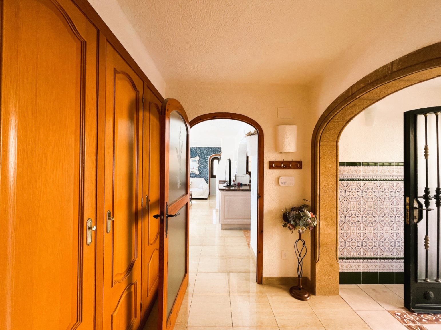 Pasillo de entrada con armarios empotrados de madera, suelos cerámicos brillantes y arcos que conectan con el salón y una zona decorada con azulejos tradicionales.