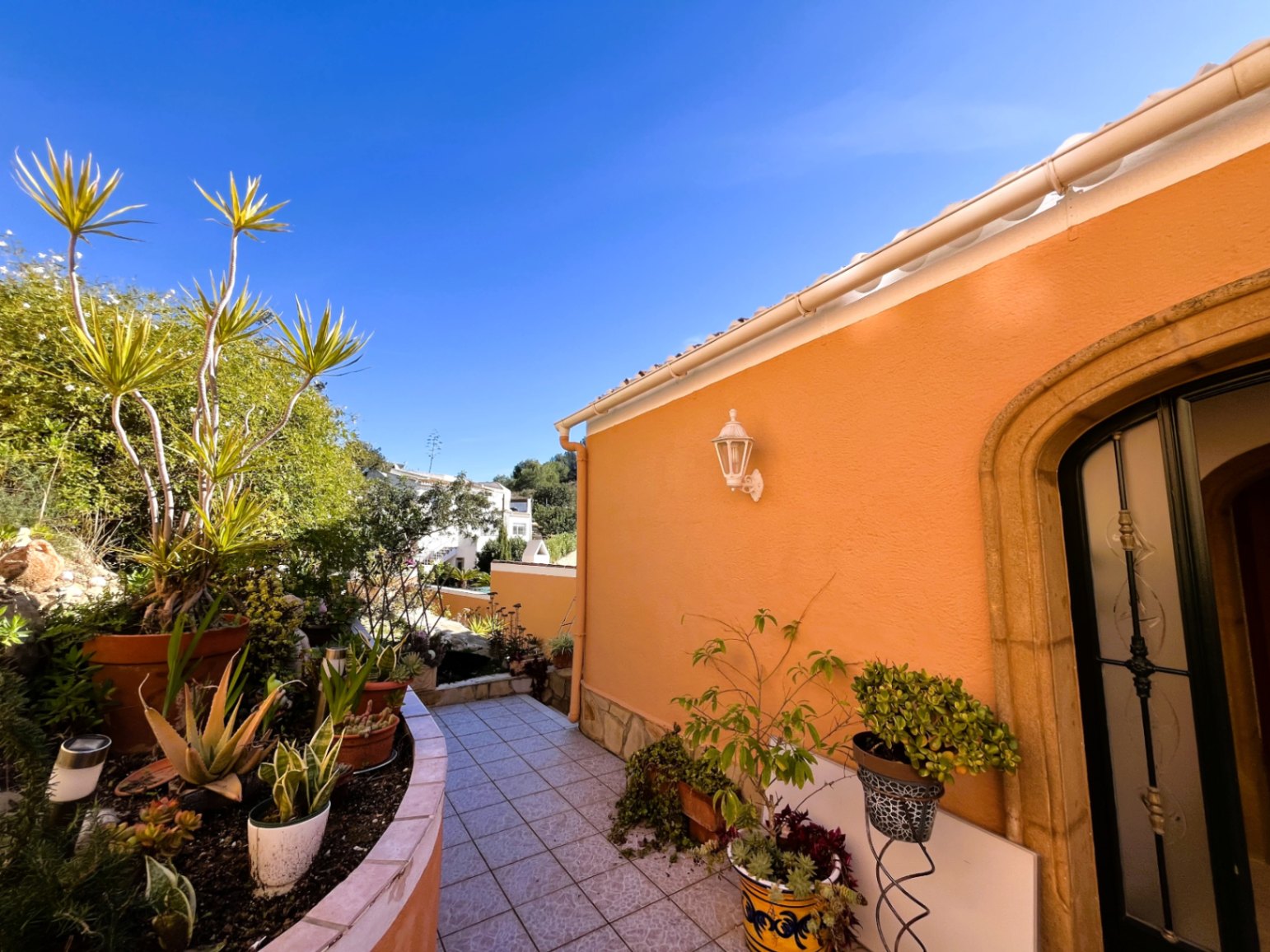 Pasillo exterior con suelo cerámico y jardinera curva, destacando la fachada ocre con arco de piedra natural y vegetación mediterránea variada.