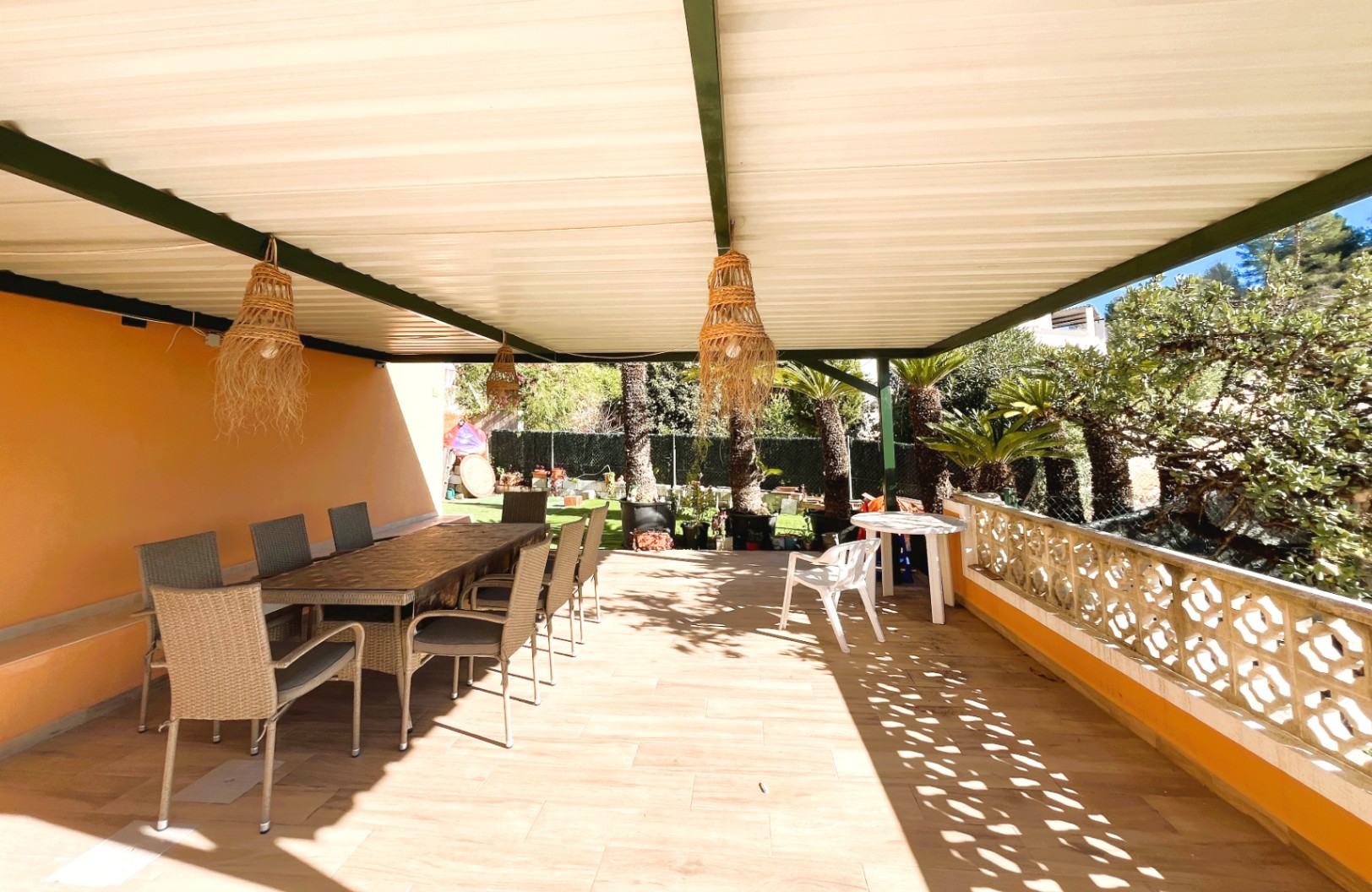 Terraza cubierta con zona de comedor exterior, lámparas de mimbre colgantes y balaustrada decorativa con vistas al jardín mediterráneo.