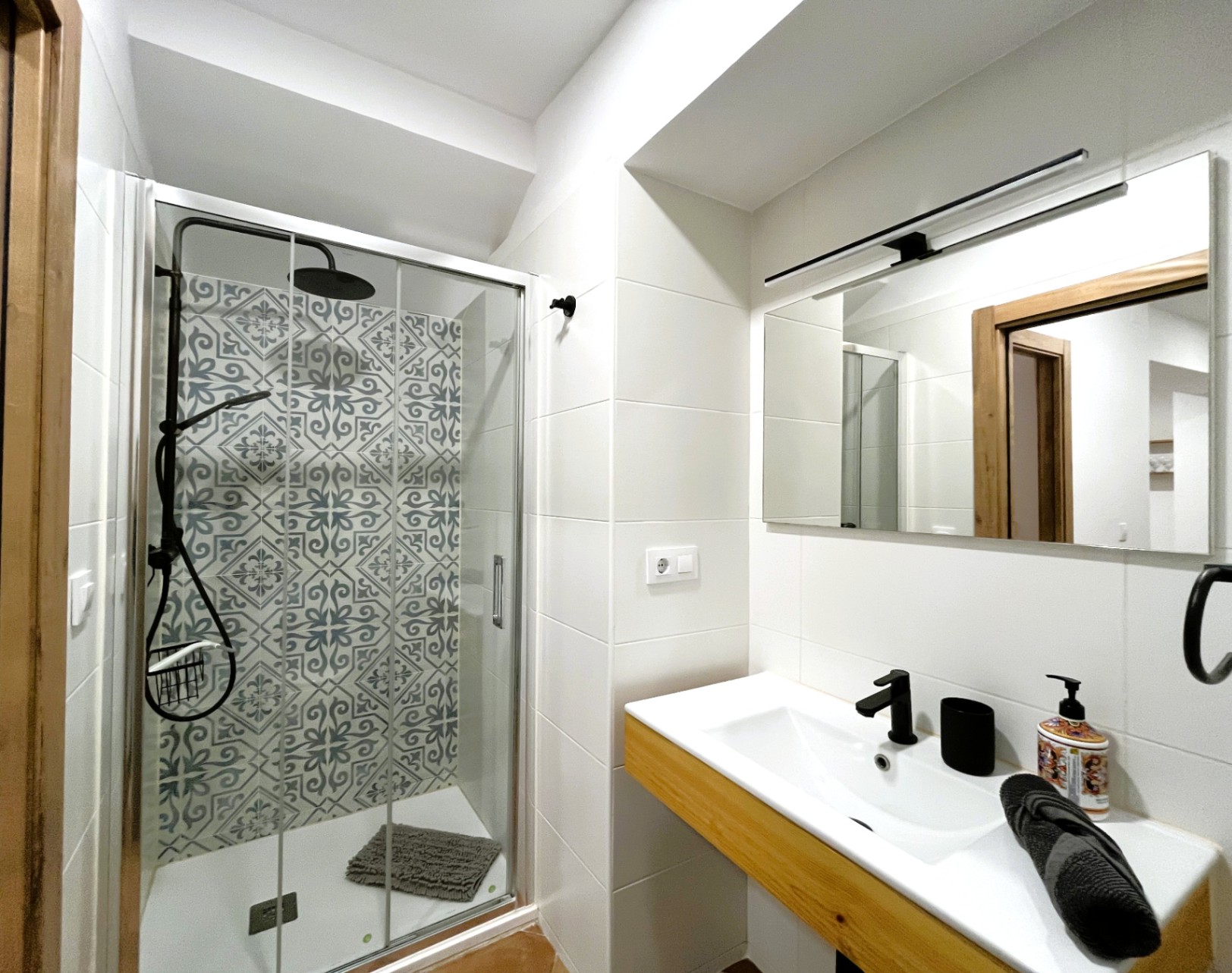 Baño moderno con ducha de obra, azulejos hidráulicos decorativos y grifería negra. Incluye lavabo integrado sobre mueble de madera y espejo con iluminación lineal.