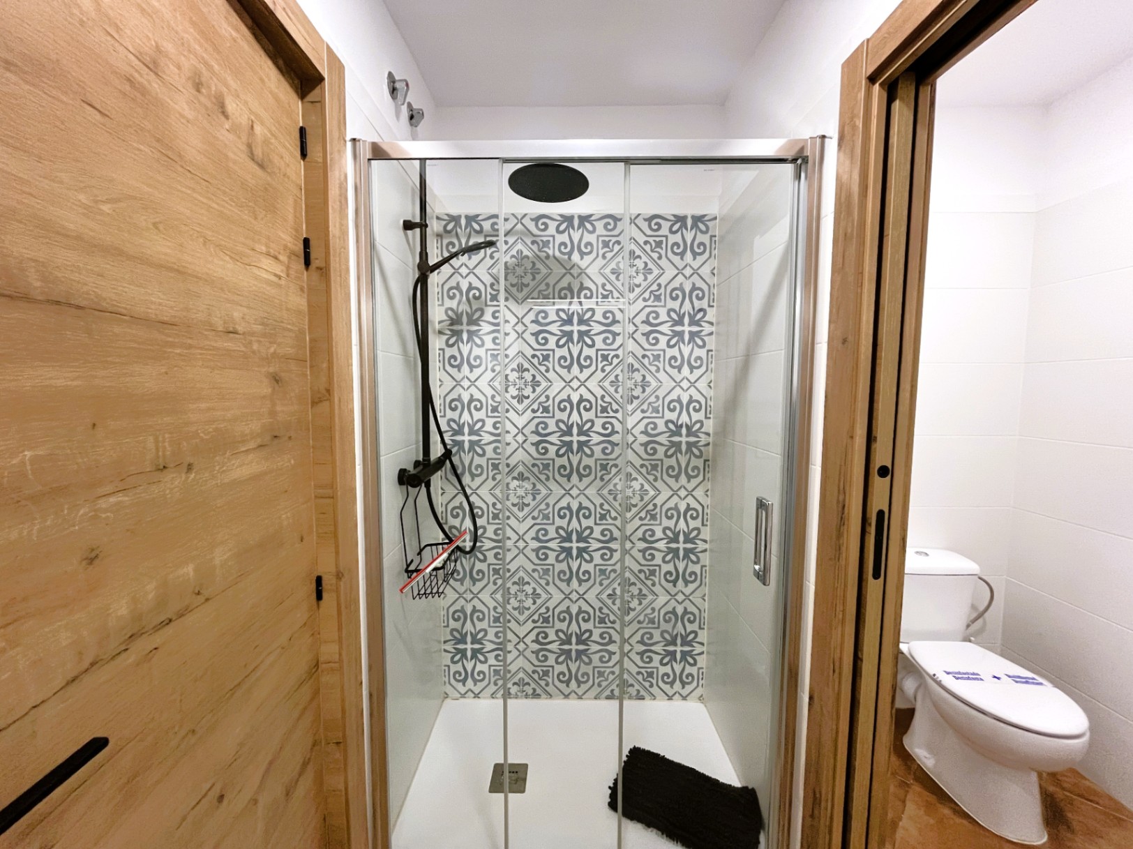 Baño reformado con ducha de obra, mampara de cristal y azulejos decorativos de estilo hidráulico. Incluye grifería negra y puertas con acabado en madera natural.