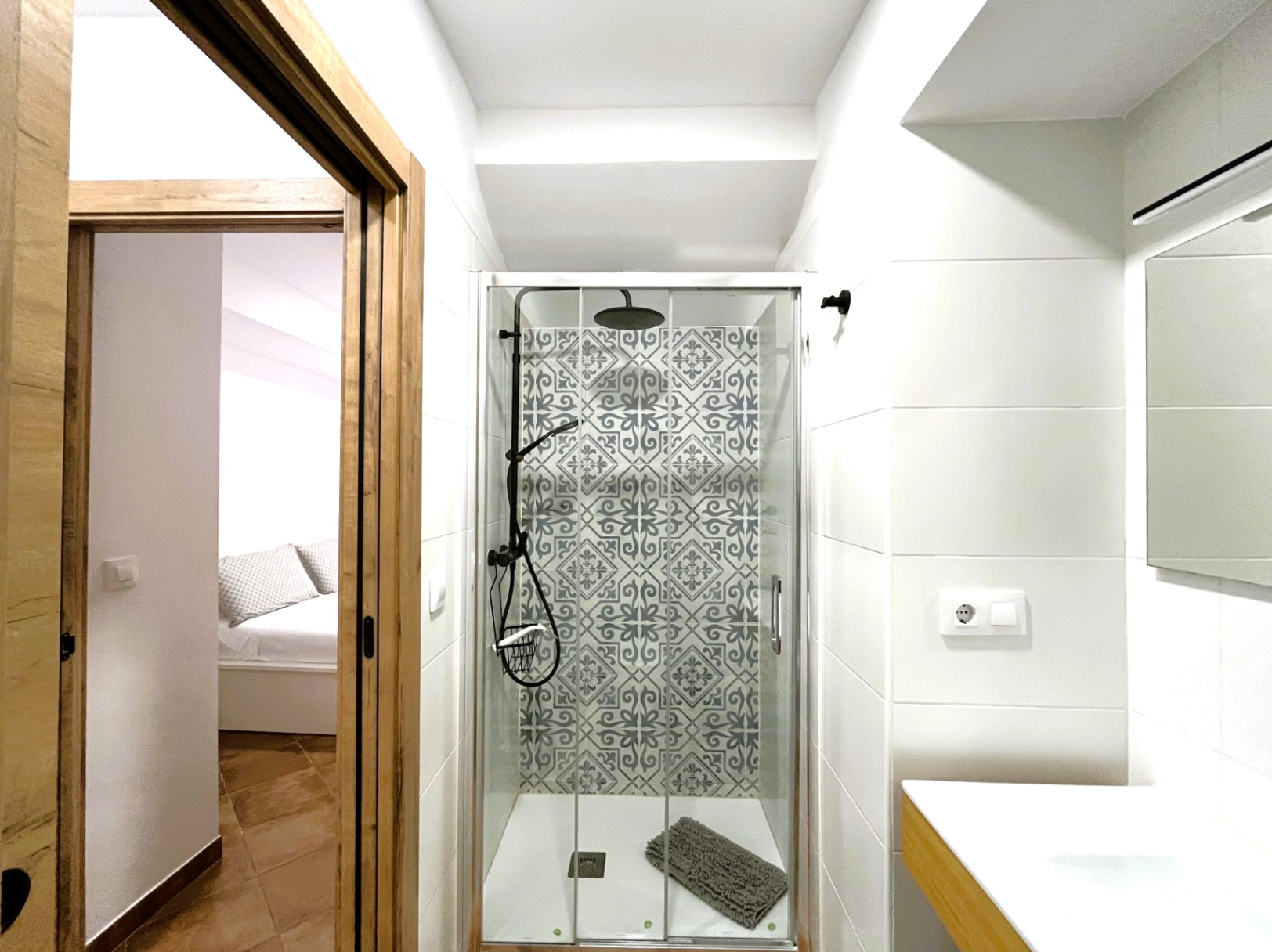 Baño reformado con ducha de obra, mampara de cristal y azulejos hidráulicos decorativos. Vista desde la entrada con marcos de madera hacia el dormitorio contiguo.