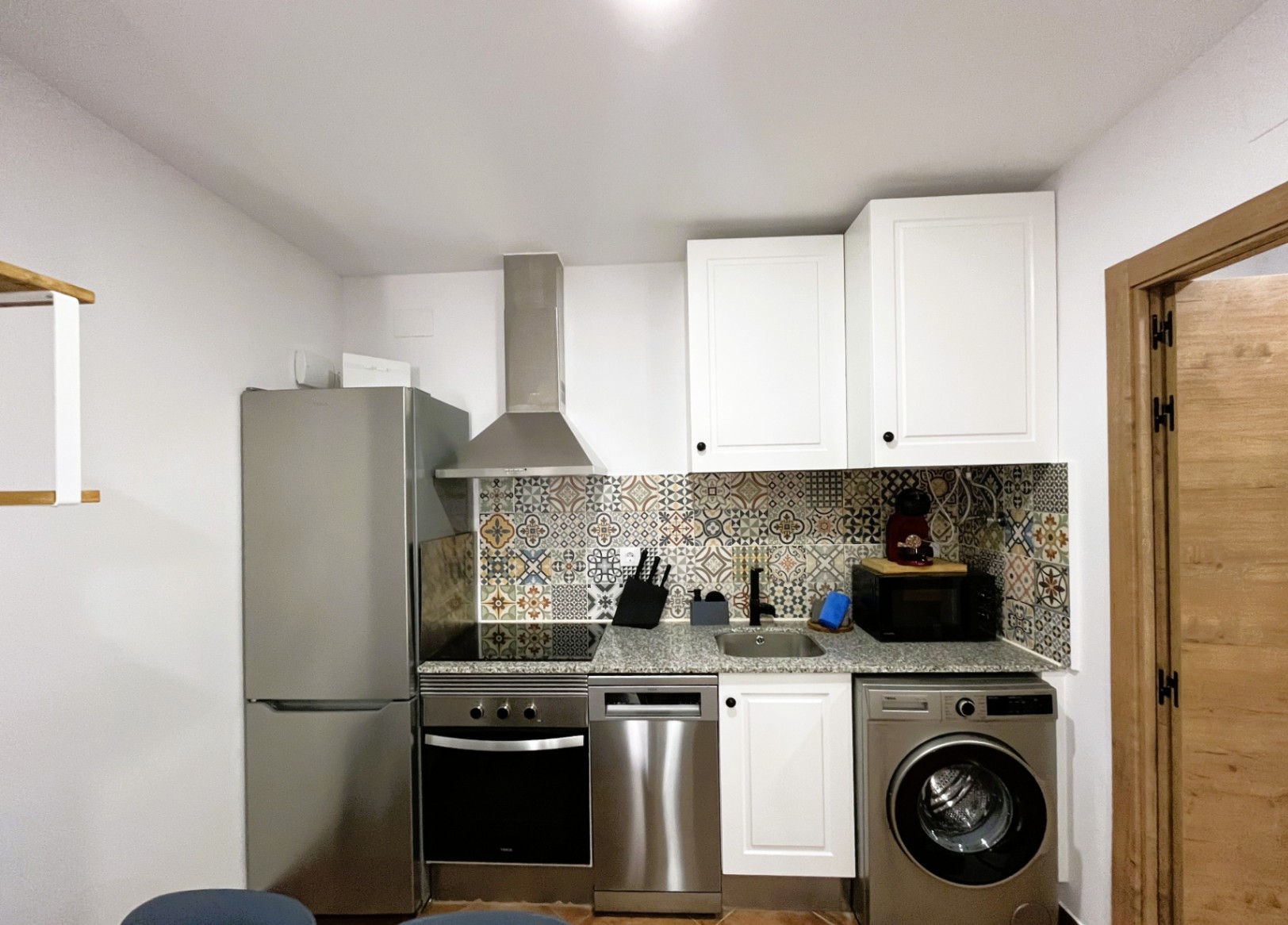 Cocina compacta reformada con encimera de granito, frontal de azulejos hidráulicos y electrodomésticos de acero inoxidable integrados, incluyendo lavadora y lavavajillas.