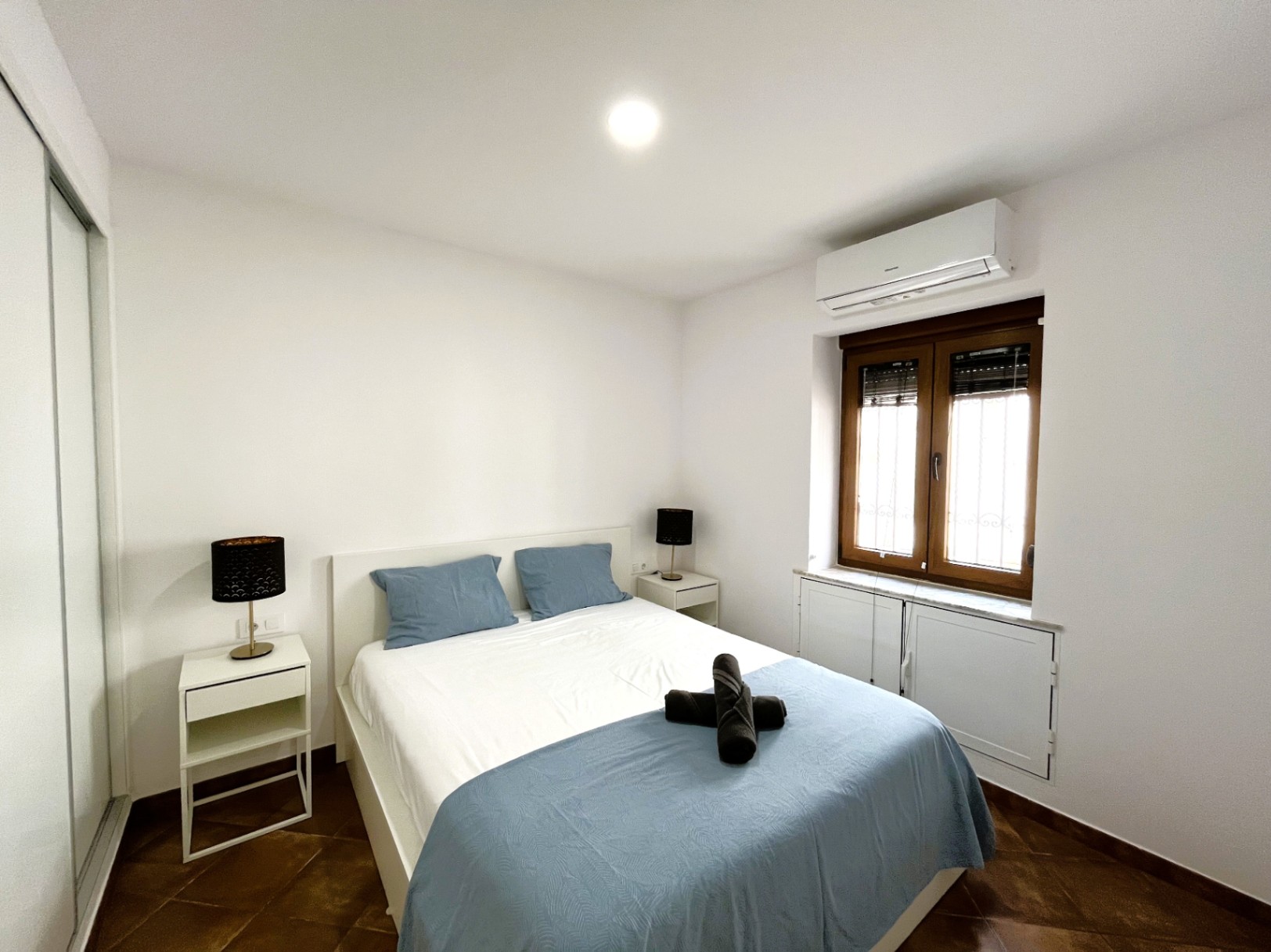 Dormitorio con cama doble, mesitas de noche blancas y aire acondicionado. Presenta suelos de baldosa rústica, armario empotrado y ventana de madera con persiana.