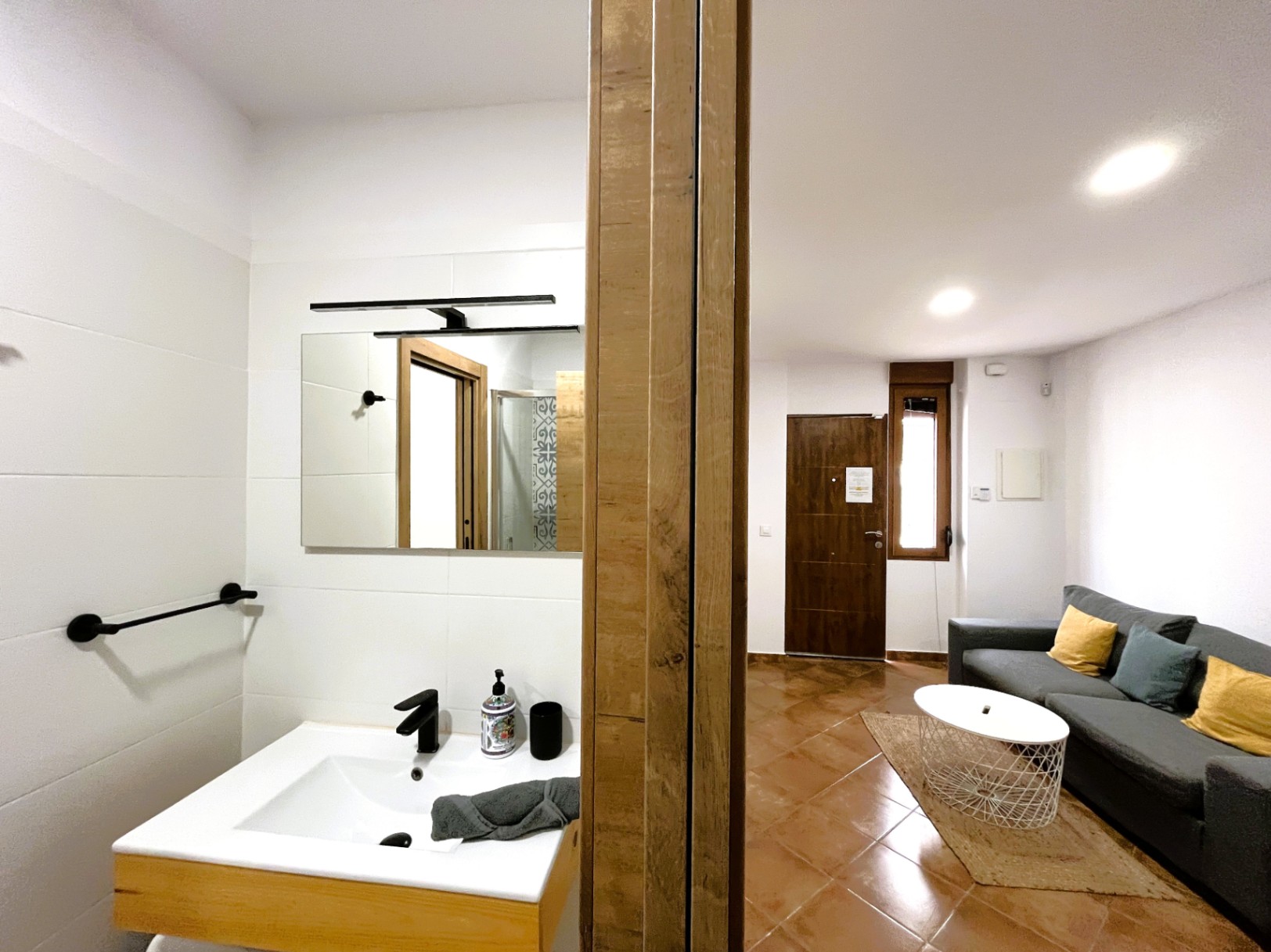 Vista de la zona de estar con suelos de terracota y sofá gris, junto a un baño moderno con lavabo de diseño, grifería negra y azulejos blancos.