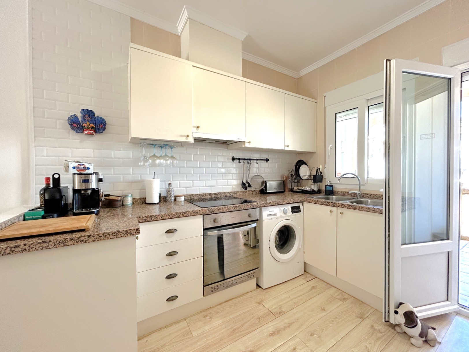 Cocina equipada con encimera de granito, mobiliario blanco y salpicadero de azulejo tipo metro. Incluye electrodomésticos integrados y suelo de gres cerámico con efecto madera.