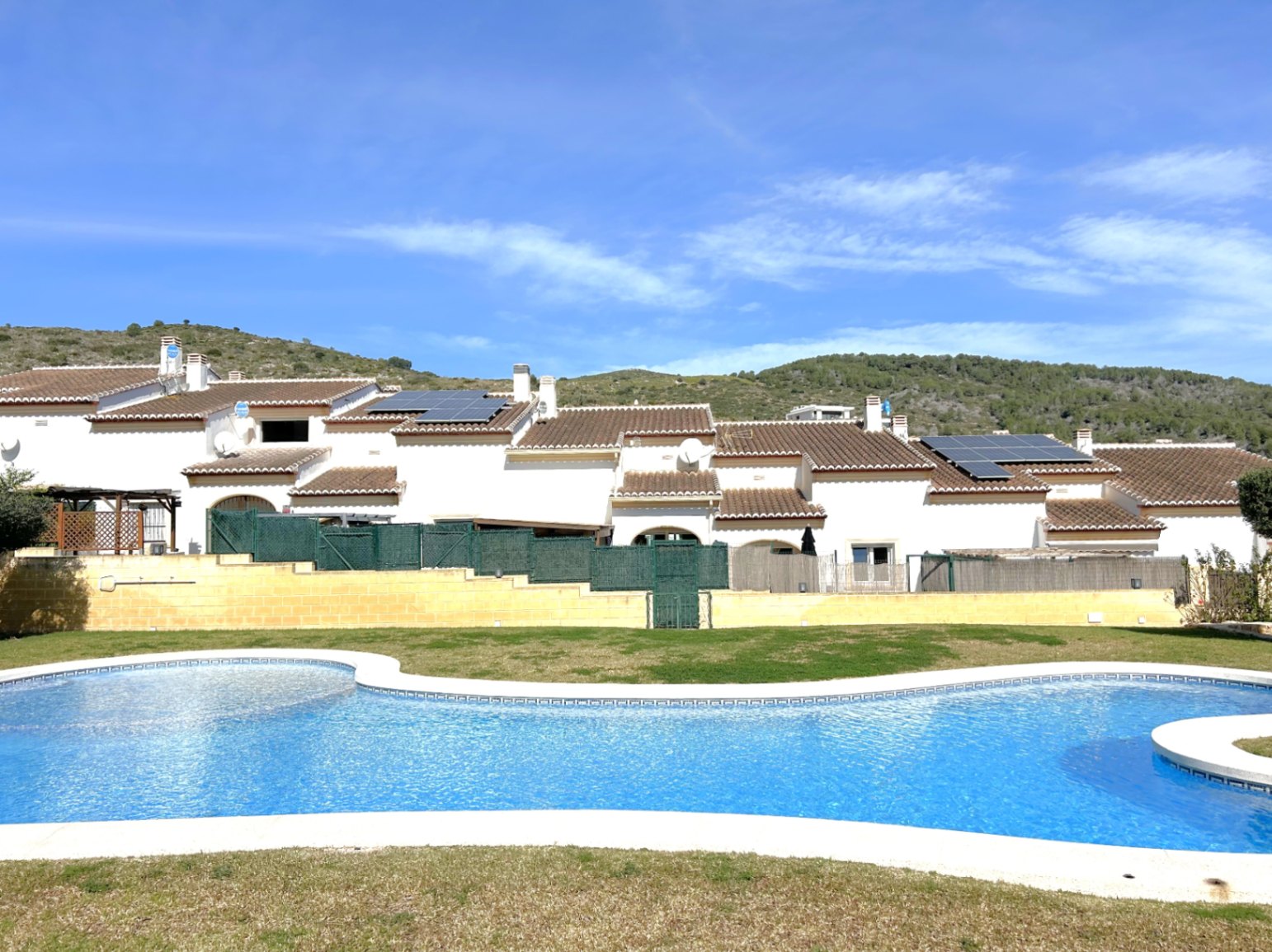 Piscina comunitaria de diseño curvo rodeada de césped, con vistas a las fachadas blancas de los adosados y el entorno natural de Benitachell.