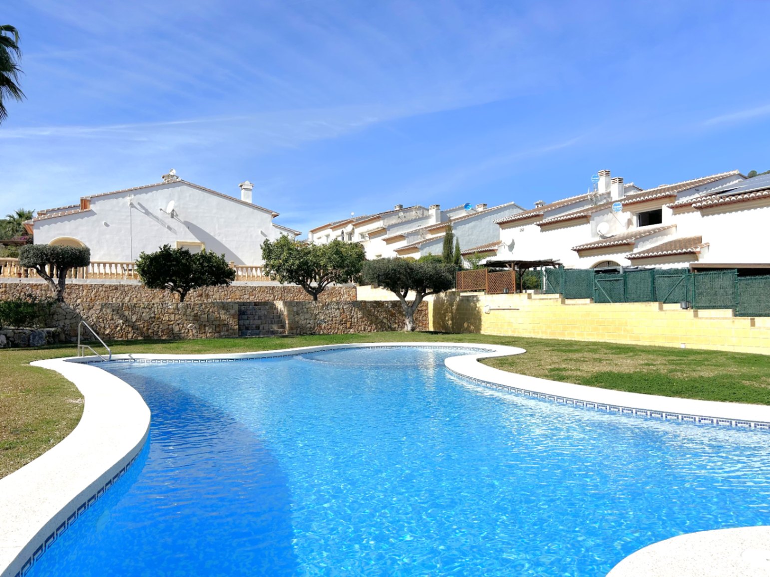 Piscina comunitaria de diseño curvo rodeada de césped y muros de piedra natural, con vistas a las viviendas blancas de estilo mediterráneo.