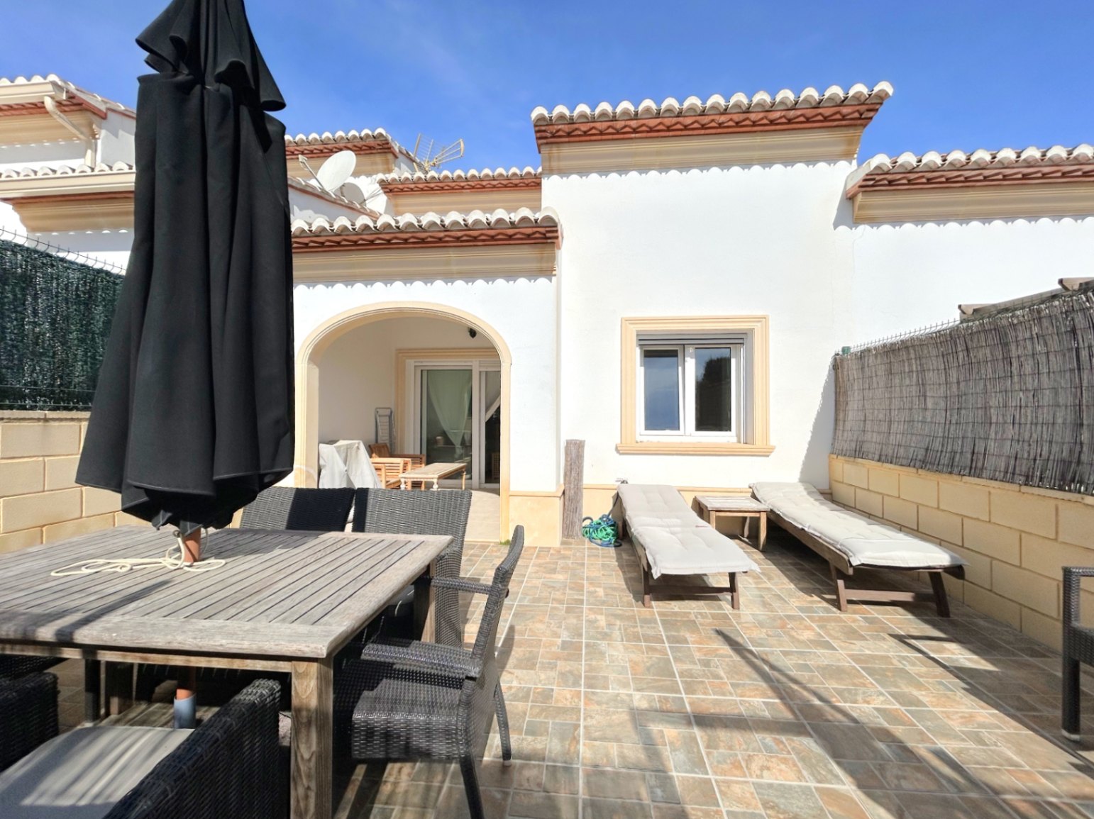 Terraza privada con suelo cerámico, zona de comedor exterior con mesa de madera y tumbonas. Incluye un porche cubierto con arco y fachada de estilo mediterráneo.
