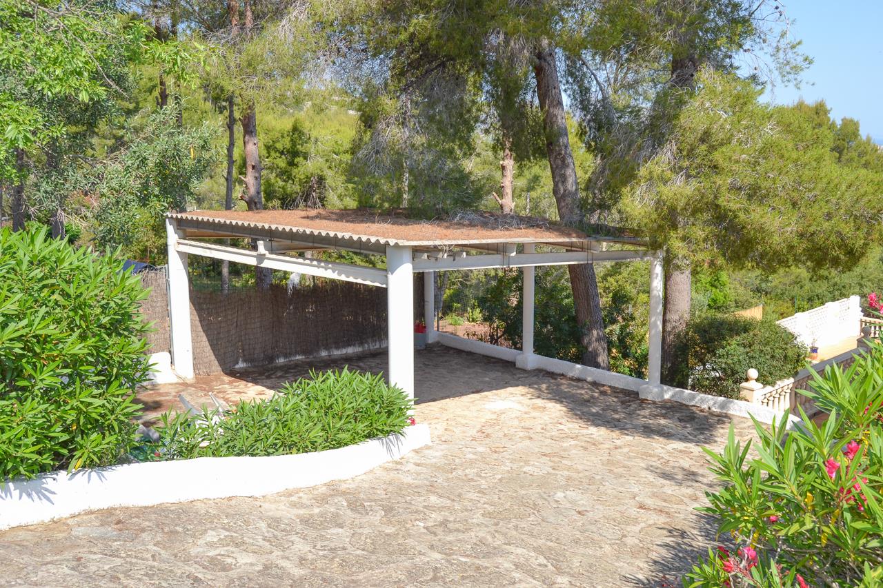 Área de aparcamiento cubierta con estructura blanca, situada junto a un camino empedrado y rodeada de pinos y vegetación mediterránea autóctona.