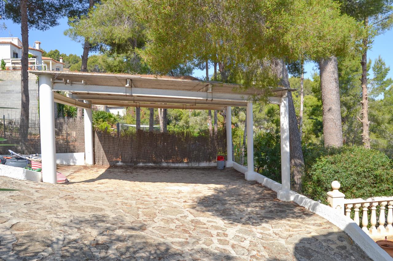 Área de aparcamiento cubierta con pilares blancos y suelo de piedra natural, integrada en el entorno natural de pinos de la parcela.