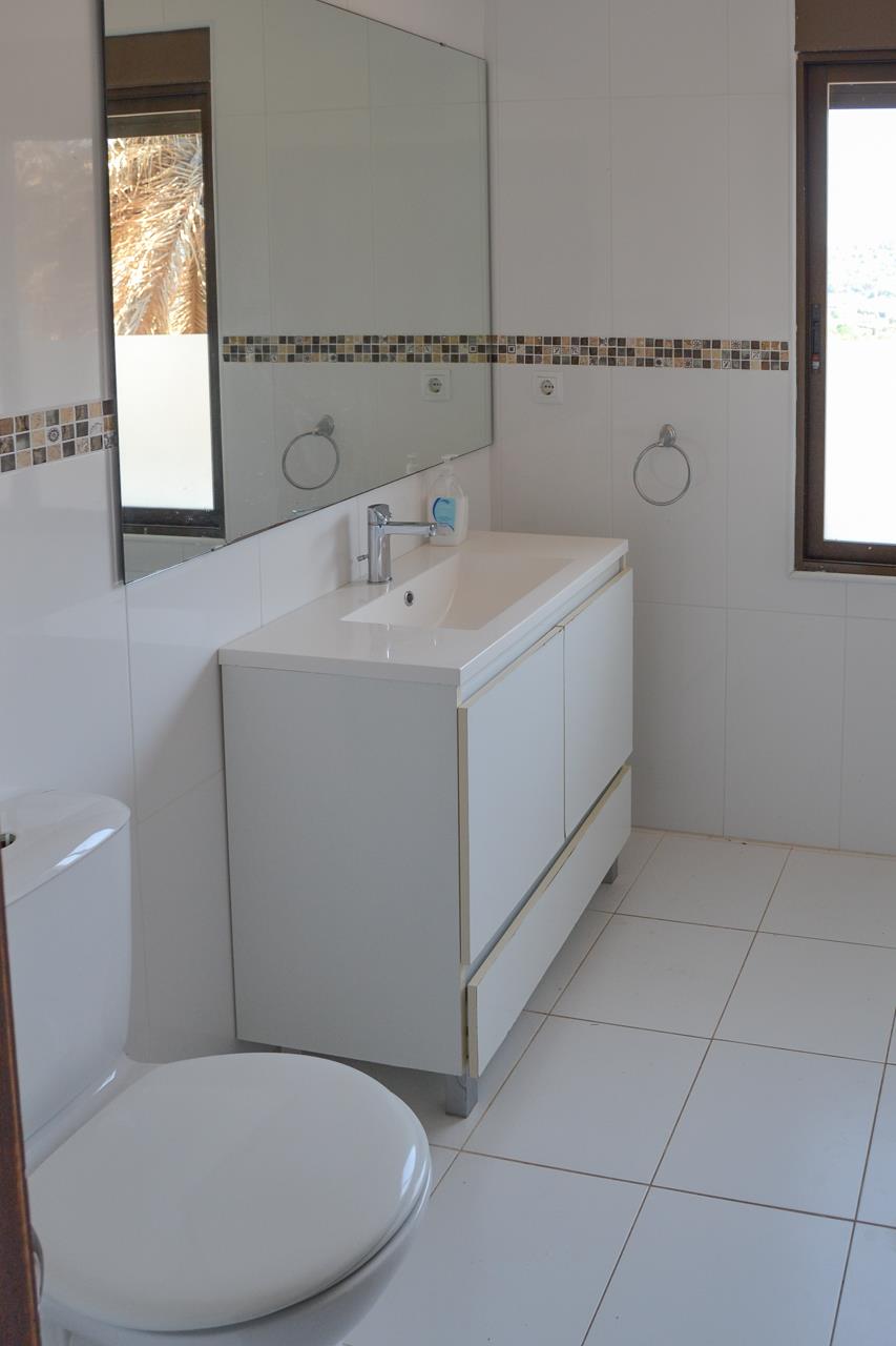 Baño con mueble de lavabo blanco, espejo de gran formato y paredes alicatadas con cenefa de mosaico. Incluye ventana con luz natural y suelo cerámico claro.