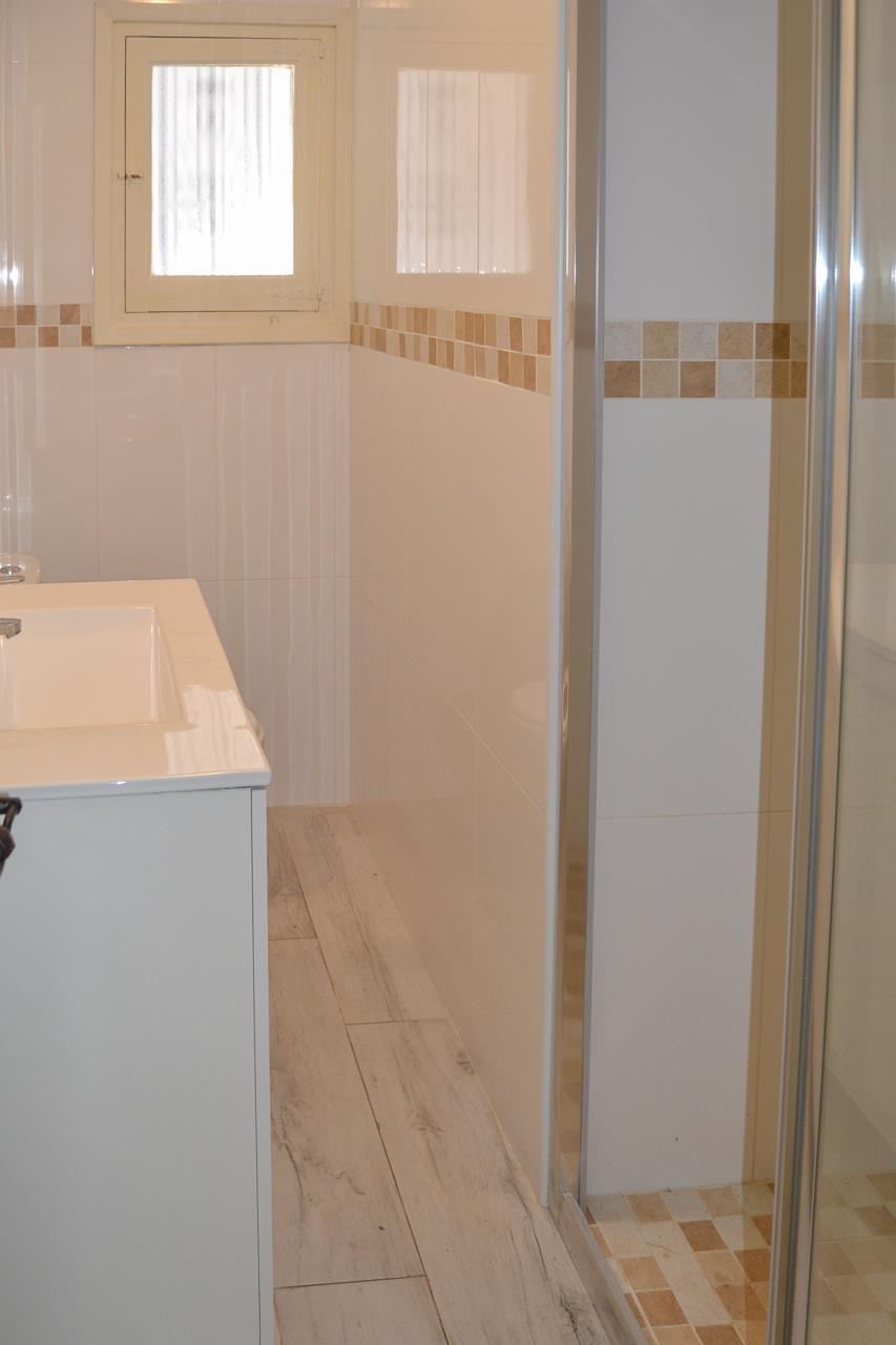 Baño moderno con mueble lavabo blanco, paredes alicatadas con cenefa decorativa y plato de ducha con mampara de cristal y suelo de mosaico.