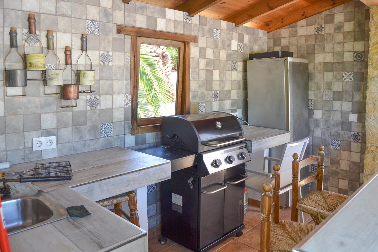 Cocina de verano equipada con barbacoa de gas, fregadero de acero inoxidable y encimeras cerámicas. Paredes decoradas con azulejos rústicos y techos con vigas de madera.