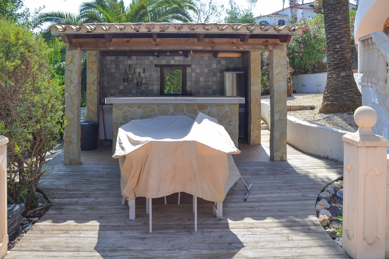 Cocina de verano exterior con barra revestida en piedra, pilares de mampostería y techado de madera sobre suelo de tarima, rodeada de vegetación mediterránea.