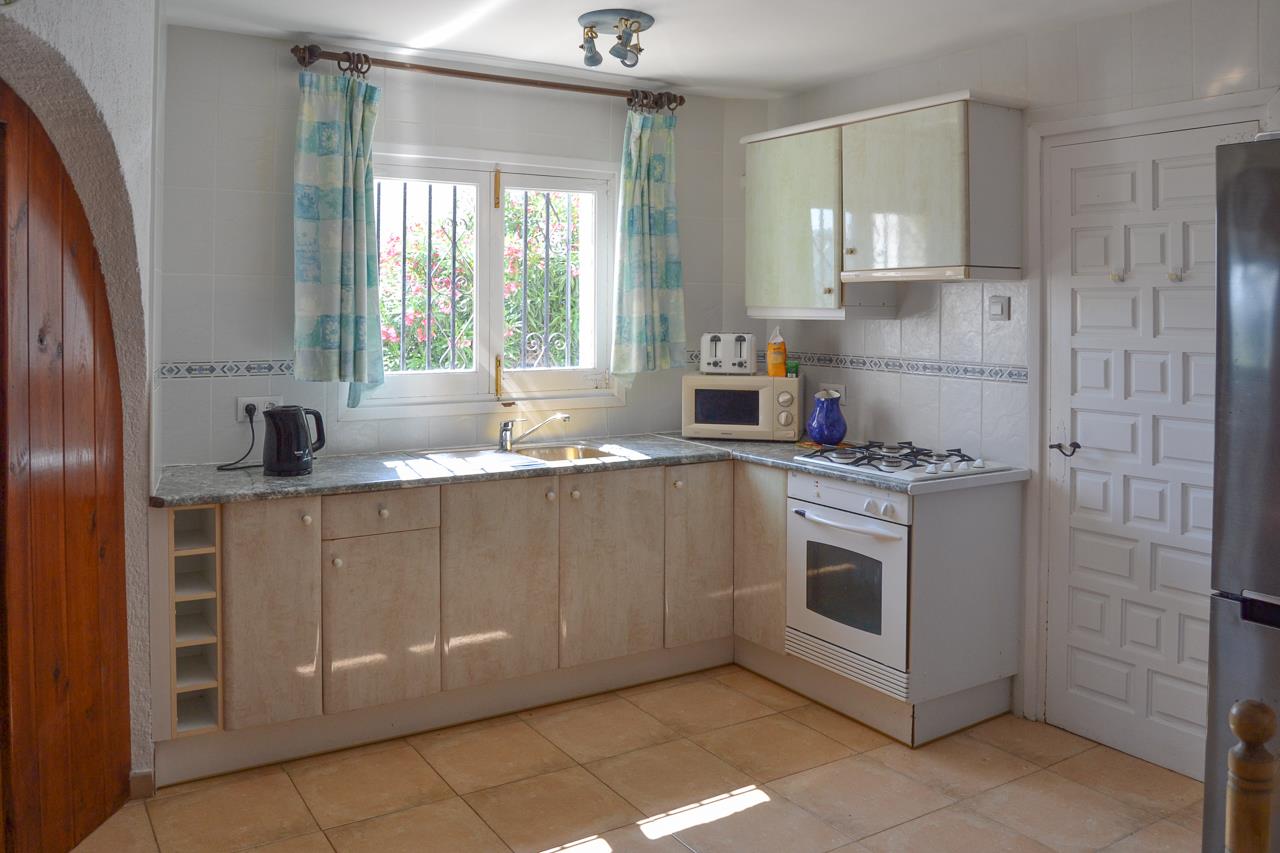 Cocina funcional con encimera de efecto granito, horno blanco y placa de gas. Incluye ventana con rejas de seguridad y vistas al exterior ajardinado.