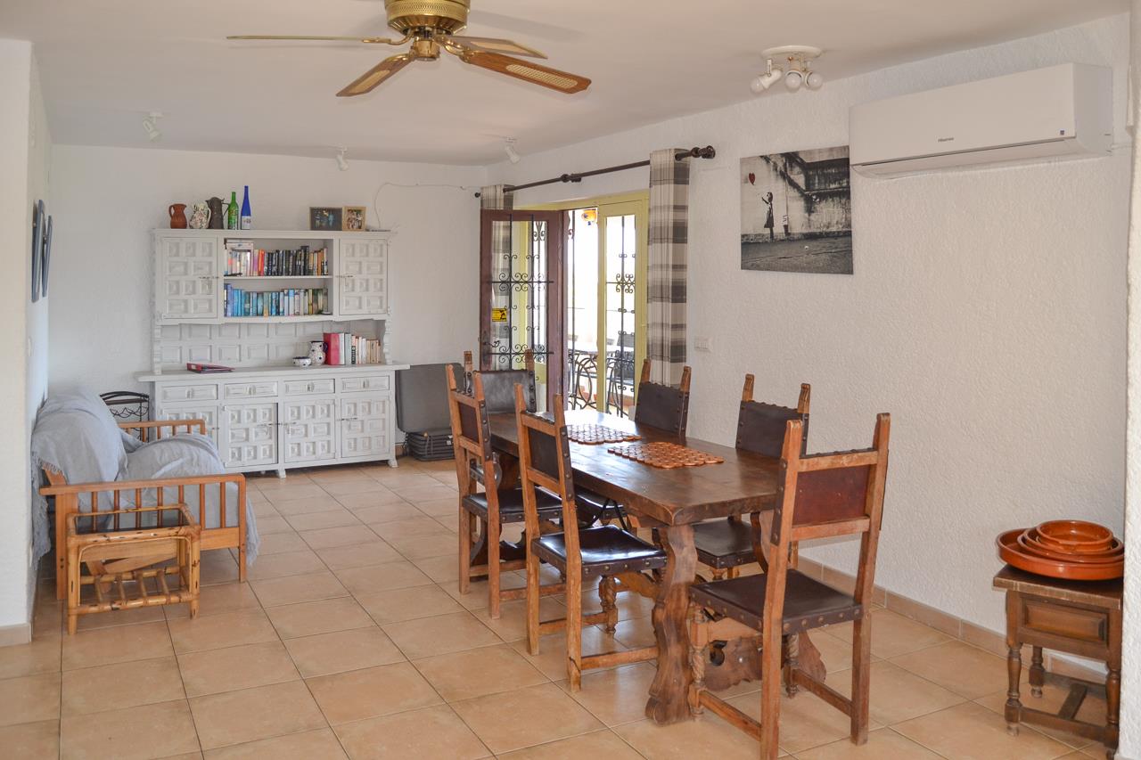 Comedor rústico con mesa de madera maciza, sillas de respaldo alto, aparador blanco y suelo de gres. Incluye aire acondicionado, ventilador de techo y acceso a la terraza.