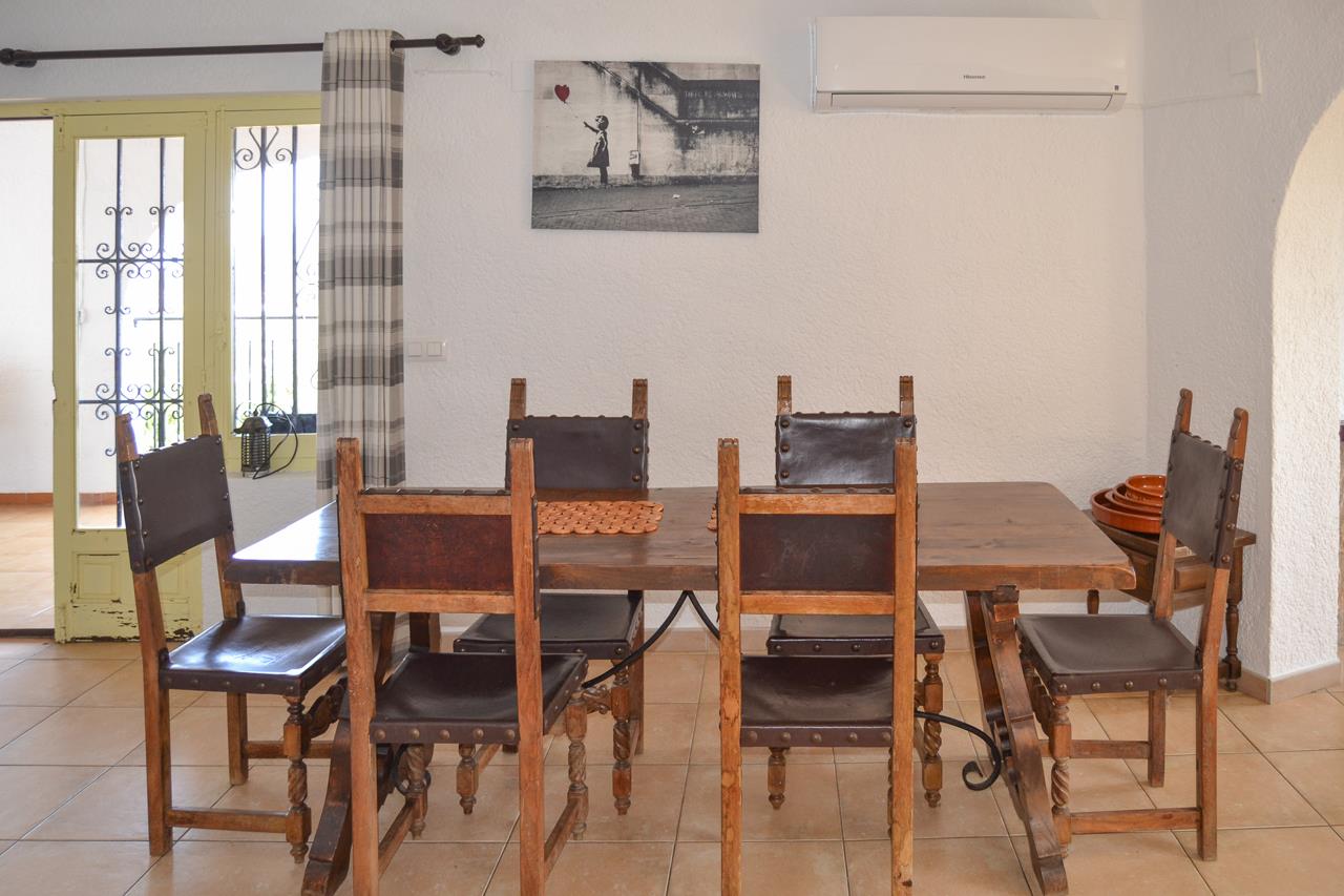 Comedor rústico con mesa de madera maciza y sillas de cuero. Incluye aire acondicionado, suelos cerámicos y acceso mediante una puerta con herrería decorativa.