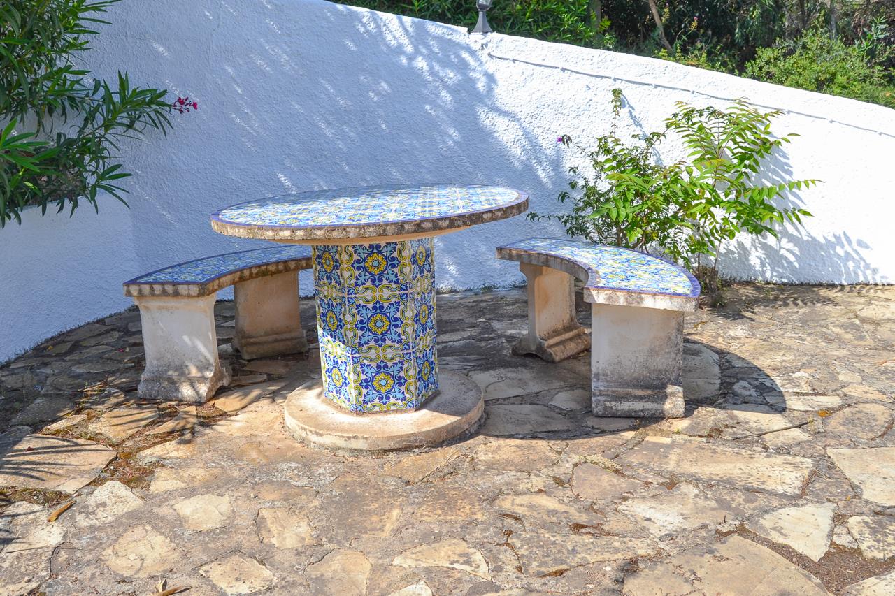 Conjunto de mesa y bancos curvos revestidos con azulejos tradicionales en tonos azules y amarillos, situados sobre un suelo de piedra natural junto a un muro blanco.