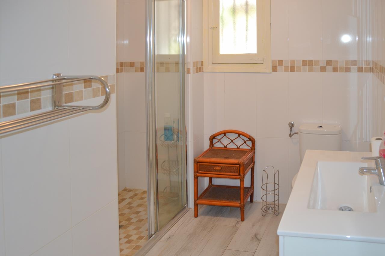 Cuarto de baño funcional con ducha de obra, mampara de cristal y mueble de lavabo blanco. Incluye azulejos claros con cenefa decorativa y luz natural.