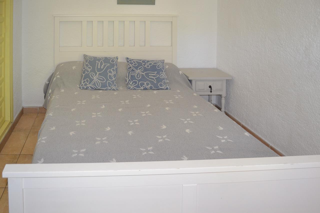 Dormitorio con cama de madera blanca, cabecero de listones y mesita de noche a juego. Cuenta con suelos de gres y paredes blancas de acabado texturizado.