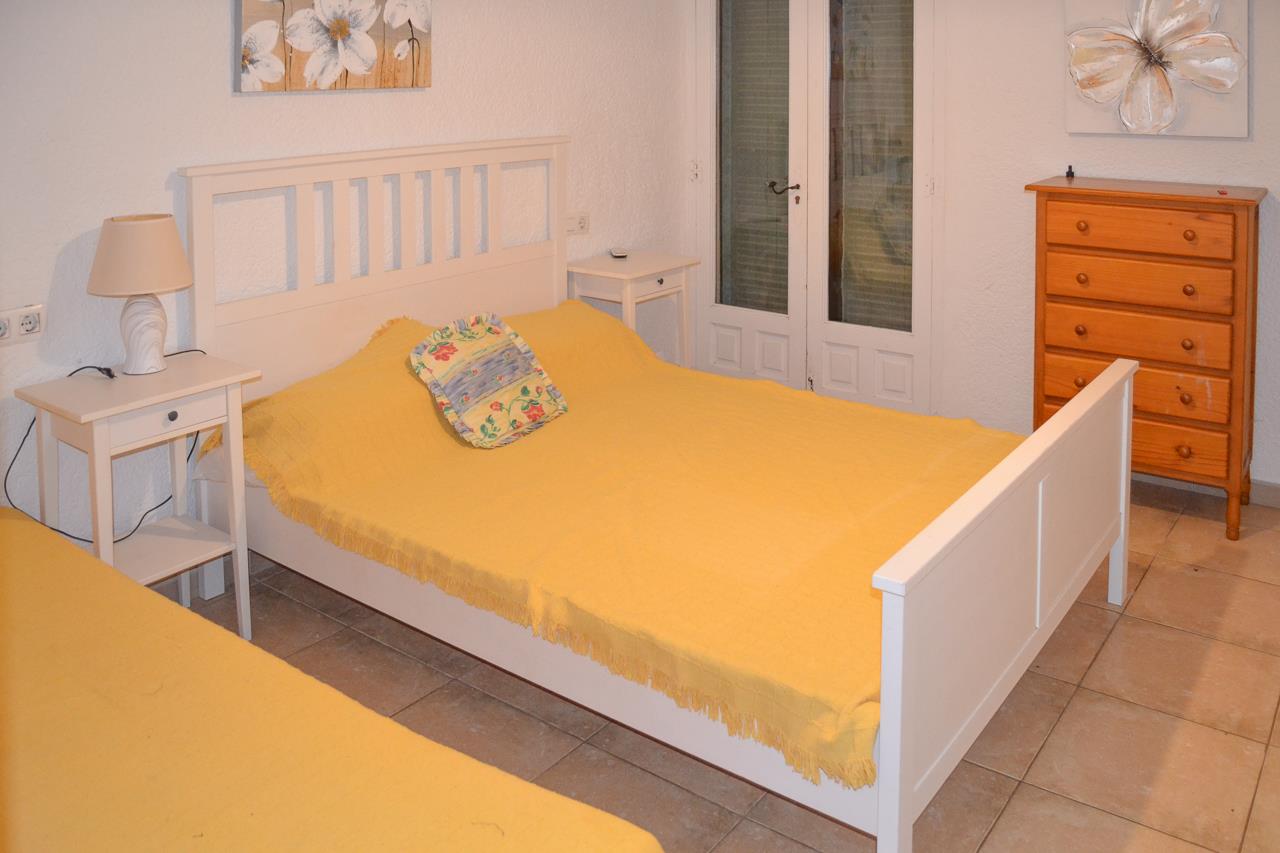Dormitorio con cama de madera blanca, mesitas de noche a juego y cómoda de madera natural sobre suelo de gres. Incluye acceso directo a través de puertas acristaladas.
