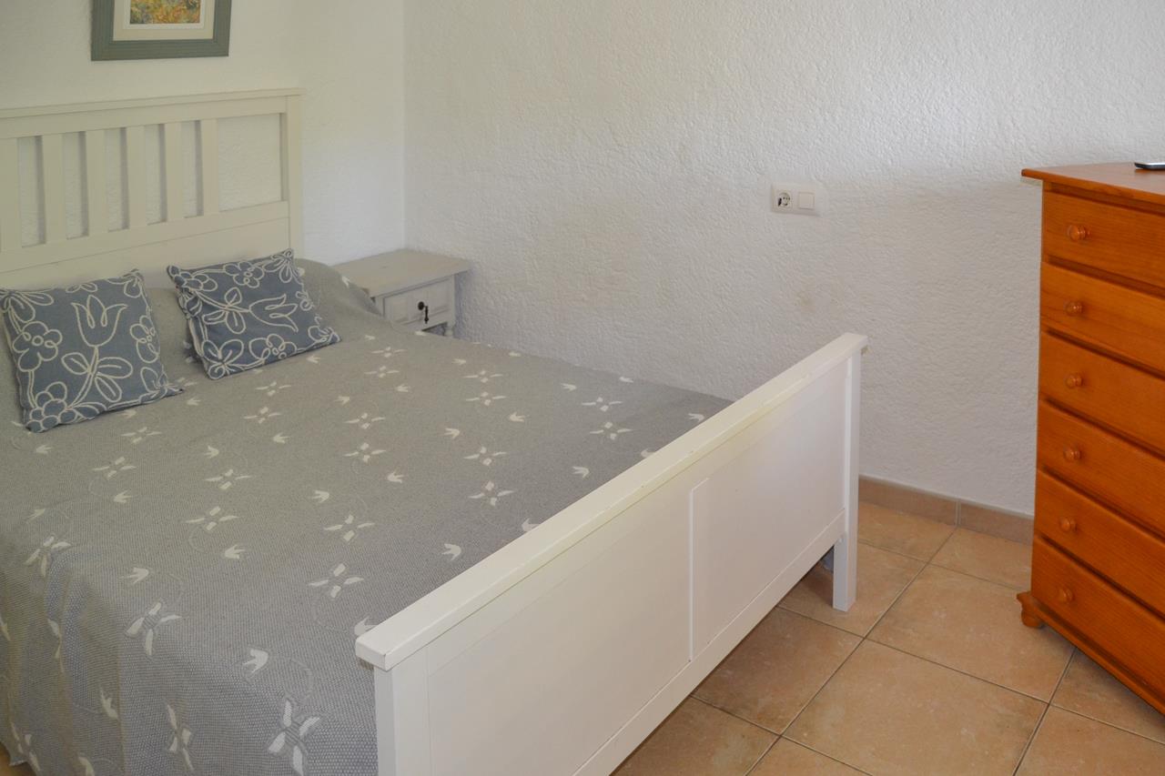 Dormitorio con cama de madera blanca, suelos de gres y una cómoda de madera natural. Las paredes blancas texturizadas aportan sencillez y luminosidad al espacio.