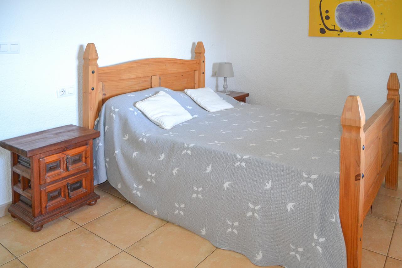 Dormitorio con cama de madera rústica, mesita de noche a juego y suelos de gres. La estancia presenta paredes blancas y luz natural.