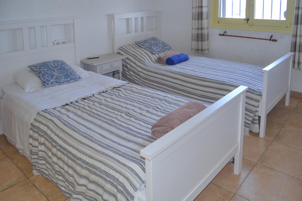 Dormitorio con dos camas individuales de madera blanca, suelos de gres y ventana con rejas de seguridad y cortinas de cuadros.