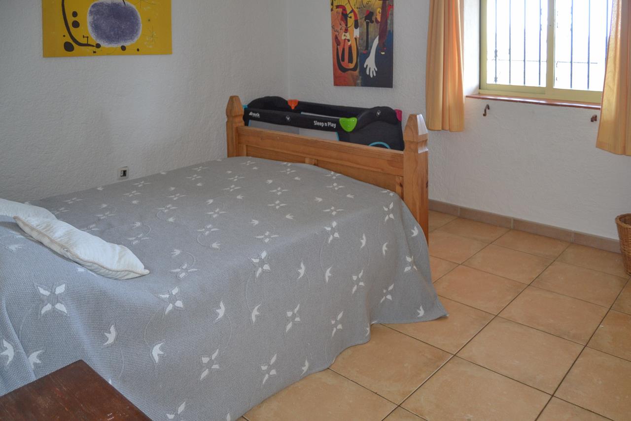 Dormitorio con suelos de gres rústico, cama de madera maciza y cuna de viaje. Dispone de ventana con rejas de seguridad y paredes blancas de acabado texturizado.