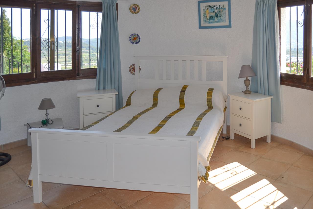 Dormitorio luminoso con cama doble de madera blanca, suelos de terracota y ventanas de madera con vistas despejadas al valle de Pedreguer.