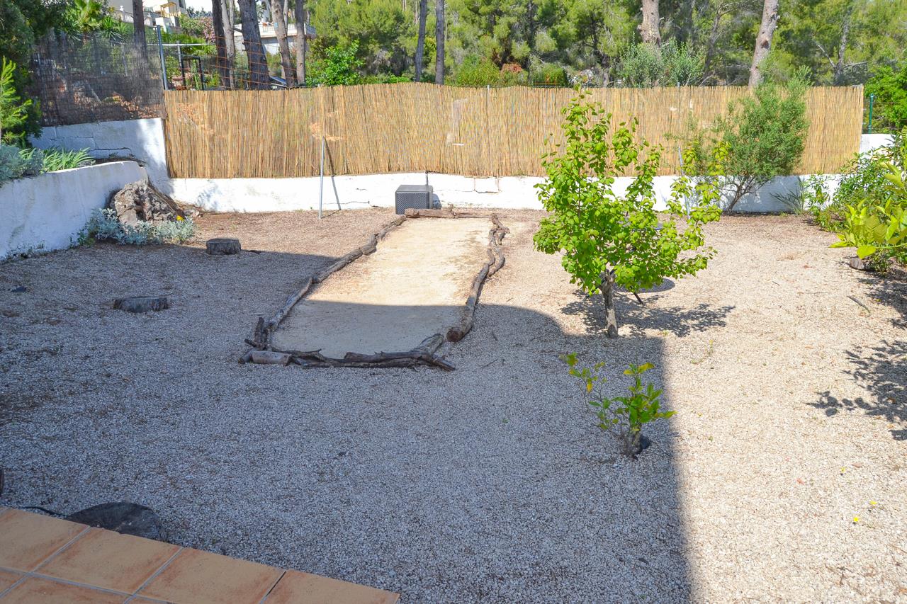 Jardín de grava con zona delimitada por troncos naturales, rodeado de vegetación mediterránea y vallado de cañizo para mayor privacidad.