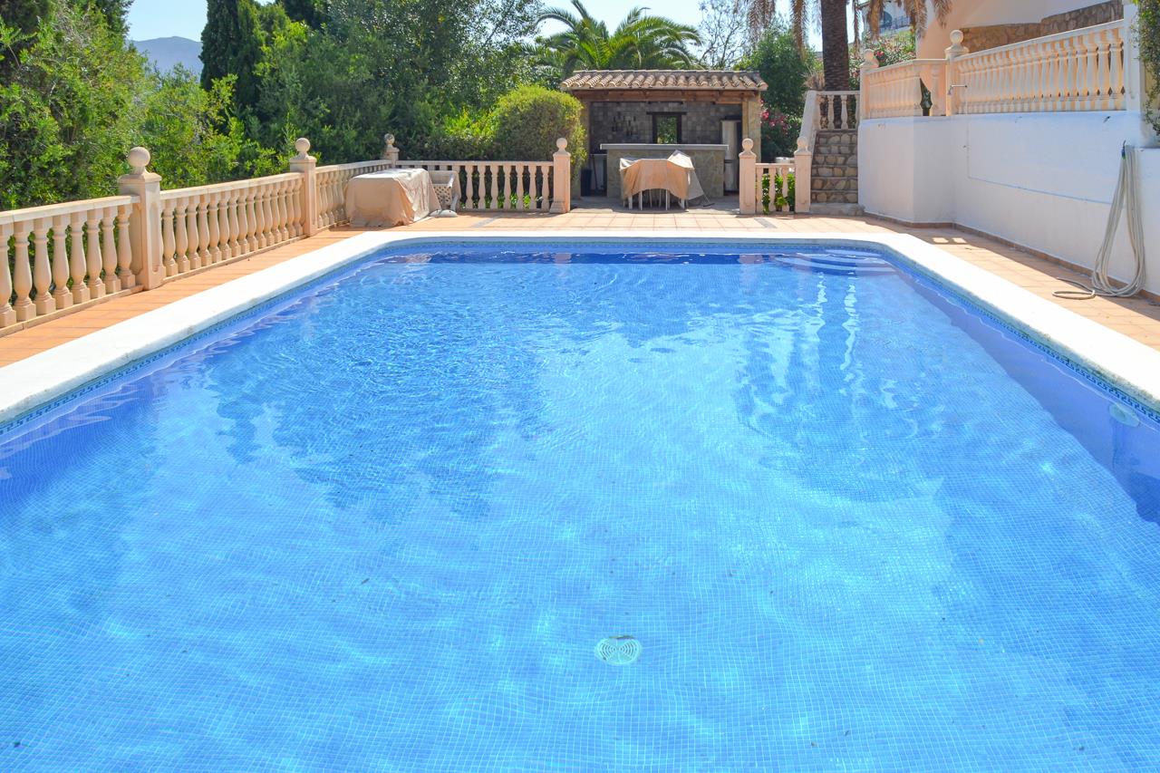 Piscina de 12x6 metros con gresite azul, rodeada de una amplia terraza con balaustrada de piedra y un quincho con cocina de verano al fondo.