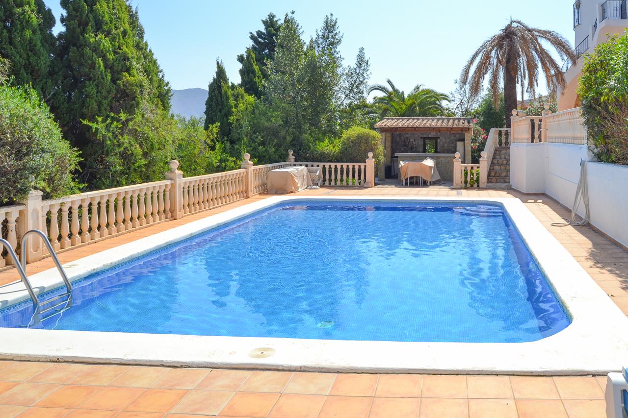 Piscina rectangular de 12x6 metros con gresite azul, rodeada de una terraza de terracota, balaustrada de piedra y cocina de verano techada con barra al fondo.