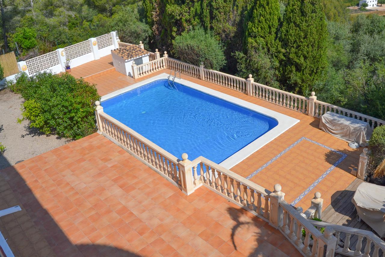 Piscina rectangular de grandes dimensiones rodeada por una terraza de baldosas de terracota y balaustrada de piedra clásica, inmersa en un entorno de vegetación mediterránea.