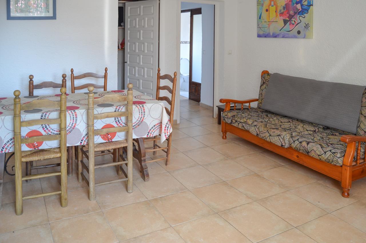 Salón comedor con suelos cerámicos, mesa de madera rústica y sofá de madera. La estancia conecta con el pasillo hacia los dormitorios y el baño.