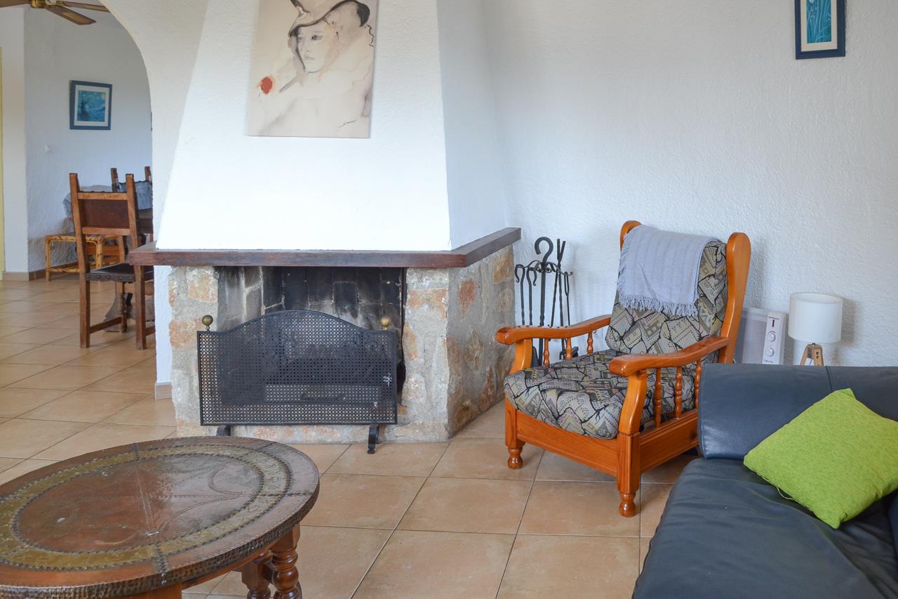 Salón con chimenea rústica de piedra y madera, suelos cerámicos y zona de estar con sillón clásico. Un arco comunica con el comedor adyacente.