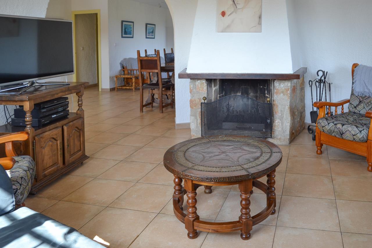 Salón rústico con chimenea de piedra y mampostería blanca, suelos de gres y mobiliario de madera tradicional que conecta con el comedor mediante un arco.