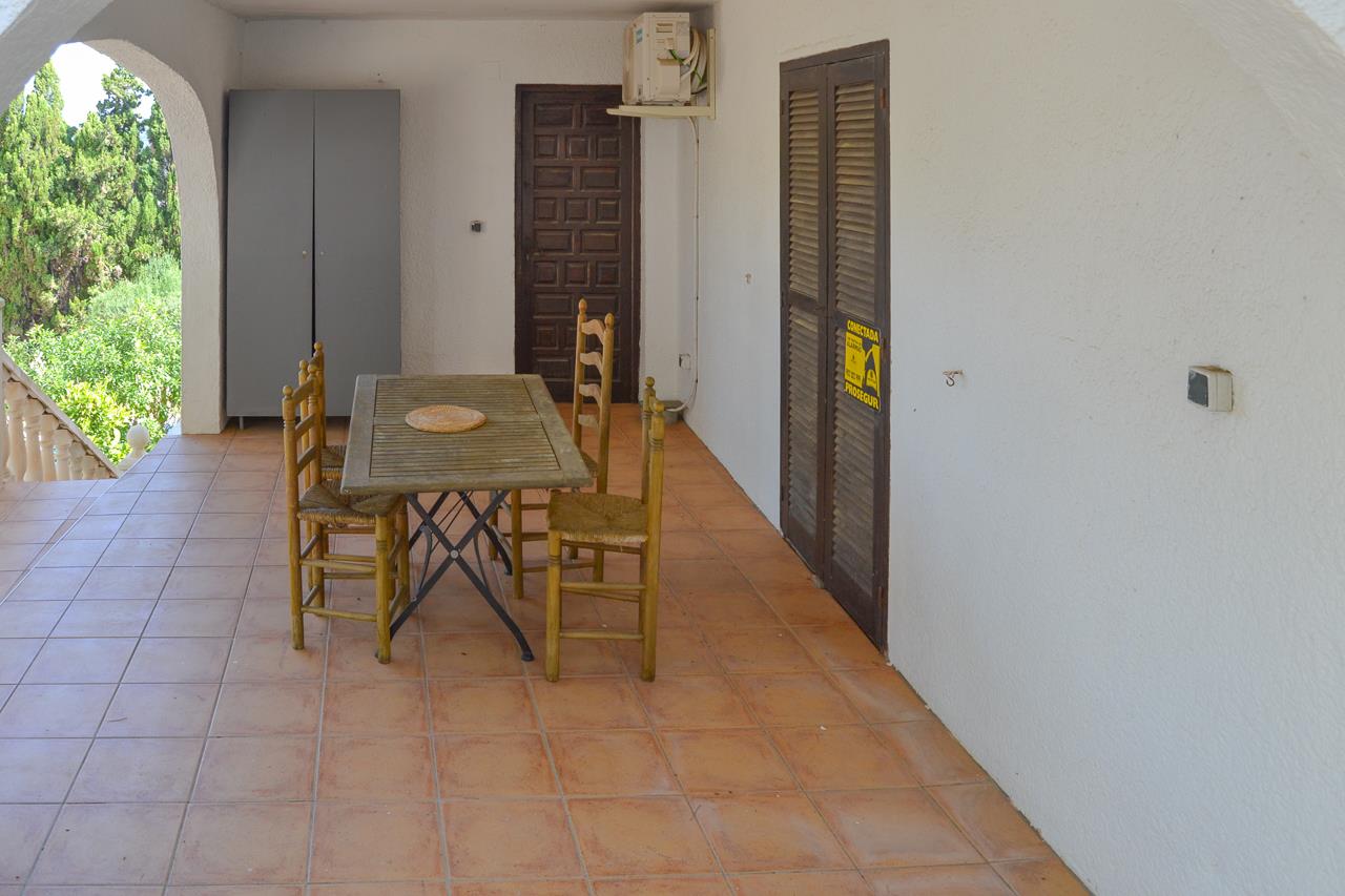 Terraza cubierta con suelos de terracota, mobiliario de comedor de madera y puertas tradicionales de madera oscura bajo una estructura de arcos blancos.