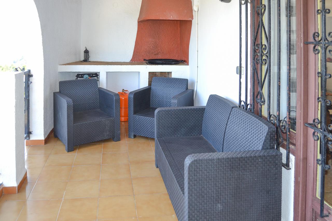 Terraza cubierta con zona de estar de ratán sintético oscuro, suelos cerámicos y barbacoa de obra integrada con campana de color terracota.