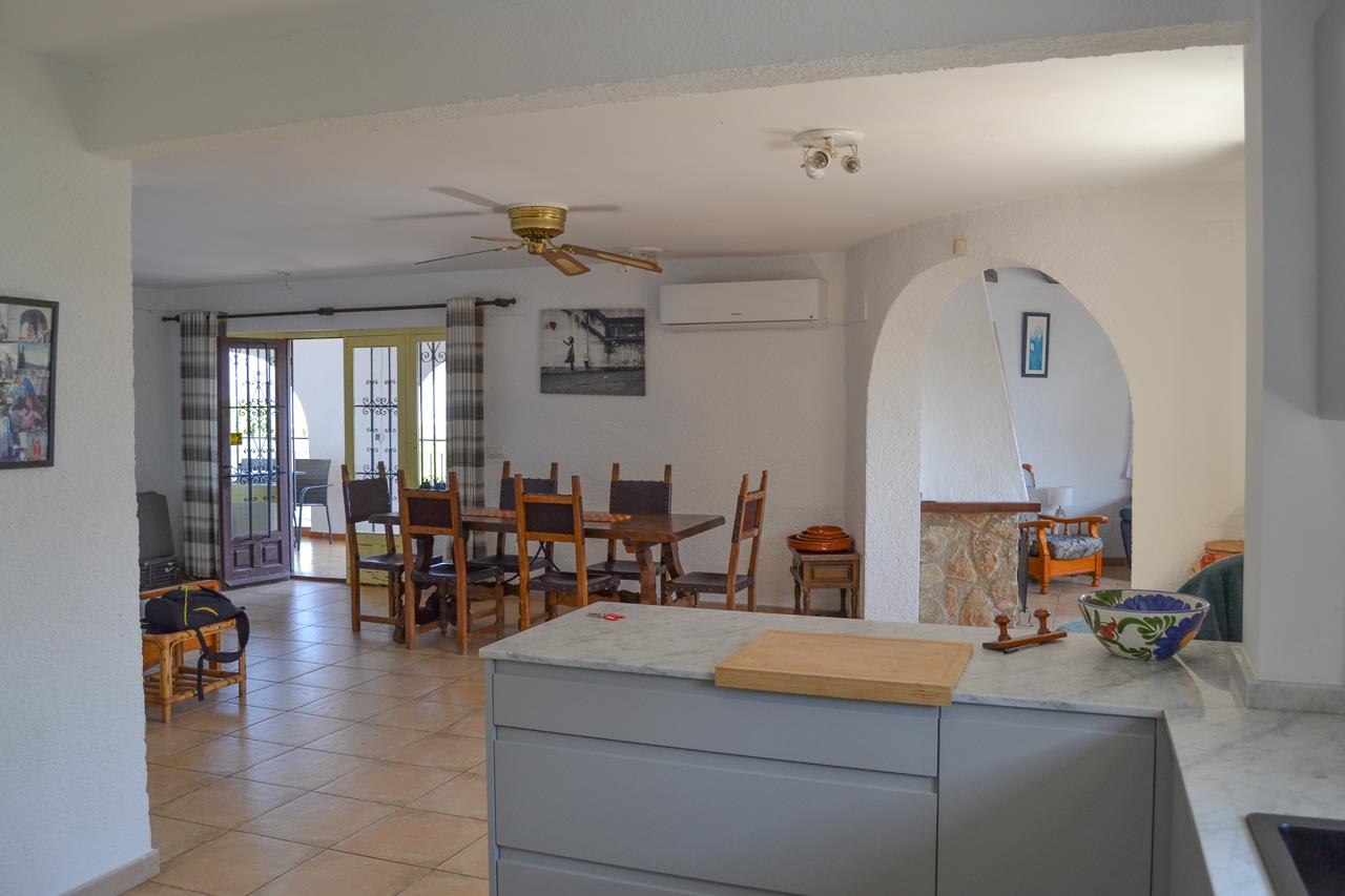 Vista desde la cocina abierta con encimera de mármol hacia el comedor. Incluye suelos cerámicos, ventilador de techo y acceso directo a la naya a través de puertas acristaladas.