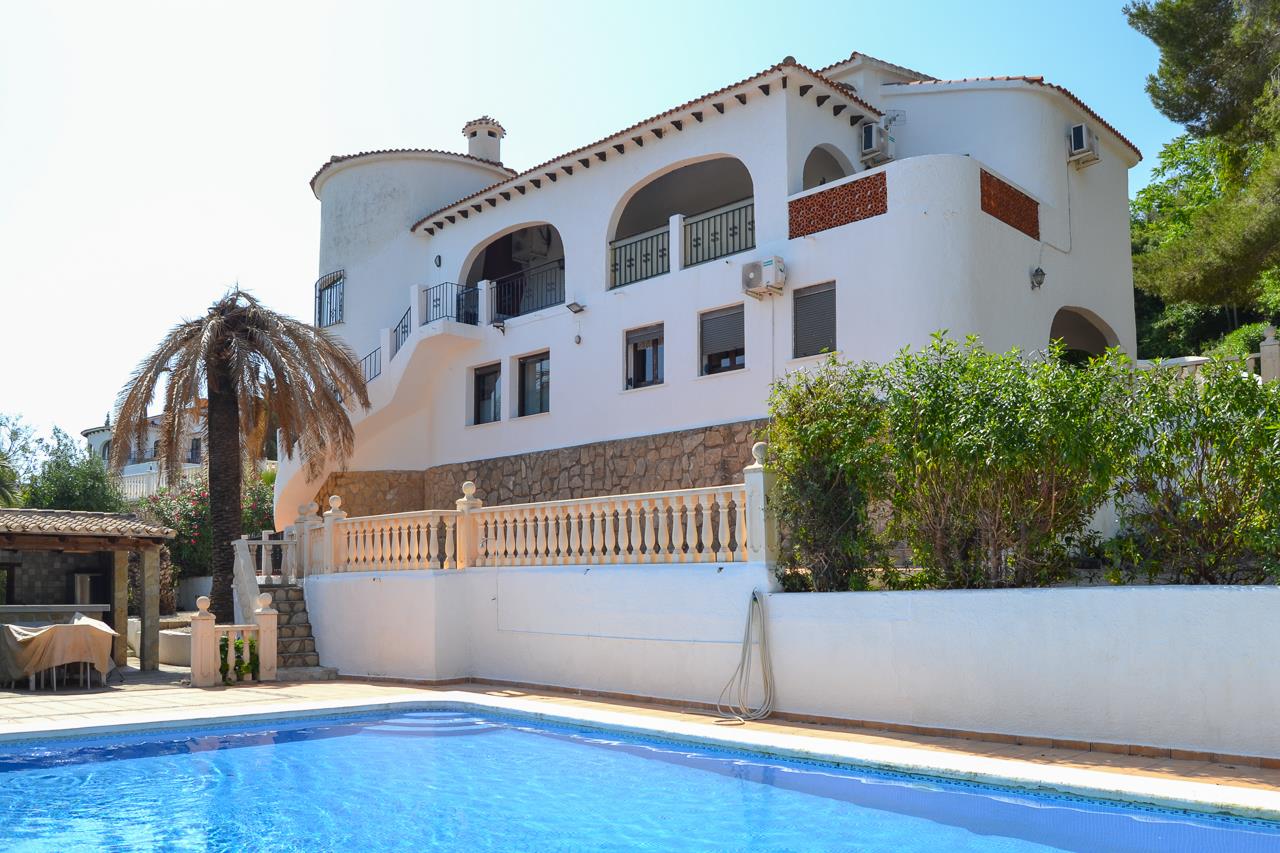 Vista exterior de la villa con piscina privada, fachadas blancas de estilo mediterráneo y terrazas cubiertas con arcos sobre un zócalo de piedra natural.