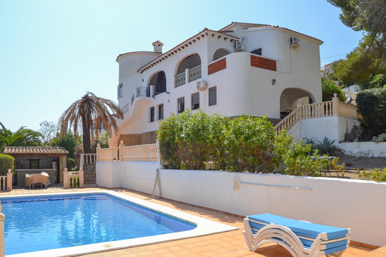 Vista exterior de la villa con piscina rectangular, amplias terrazas soleadas y fachada blanca de estilo mediterráneo con arcos y detalles en piedra.