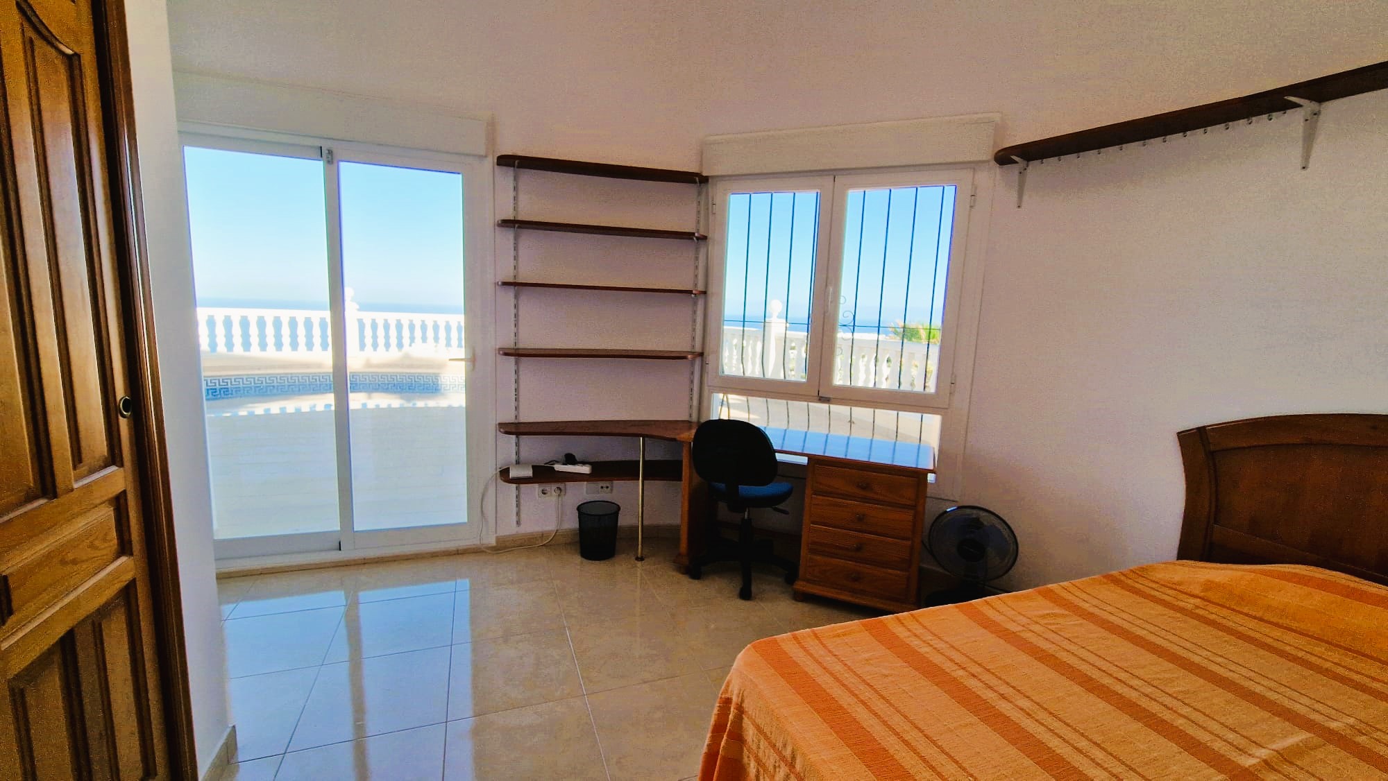 Dormitorio luminoso con escritorio de madera, estanterías y vistas al balcón con barandilla blanca.