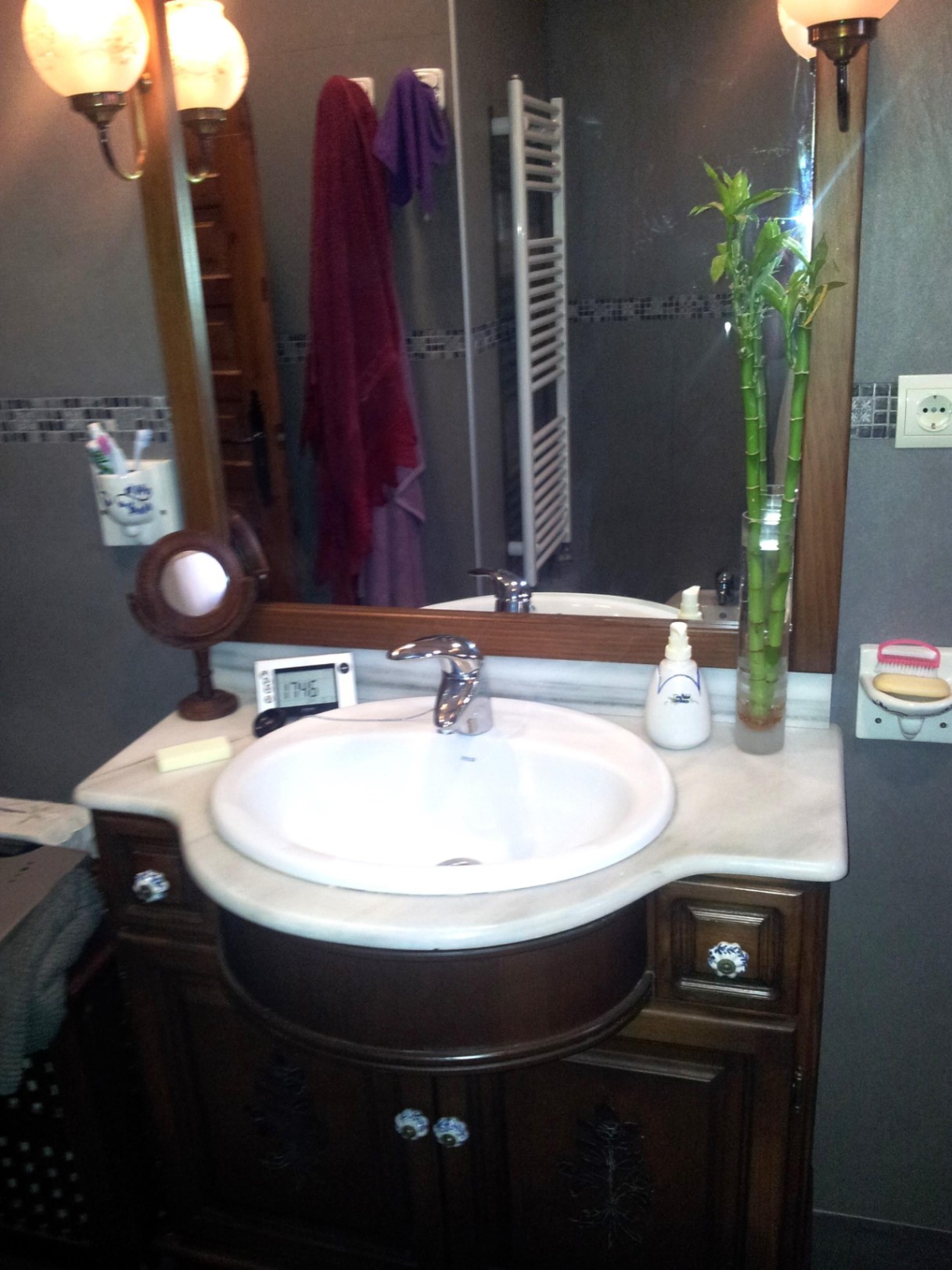 Baño con lavabo encastrado en encimera de mármol blanco, mueble de madera tallada con pomos cerámicos y un gran espejo con marco de madera y apliques clásicos.