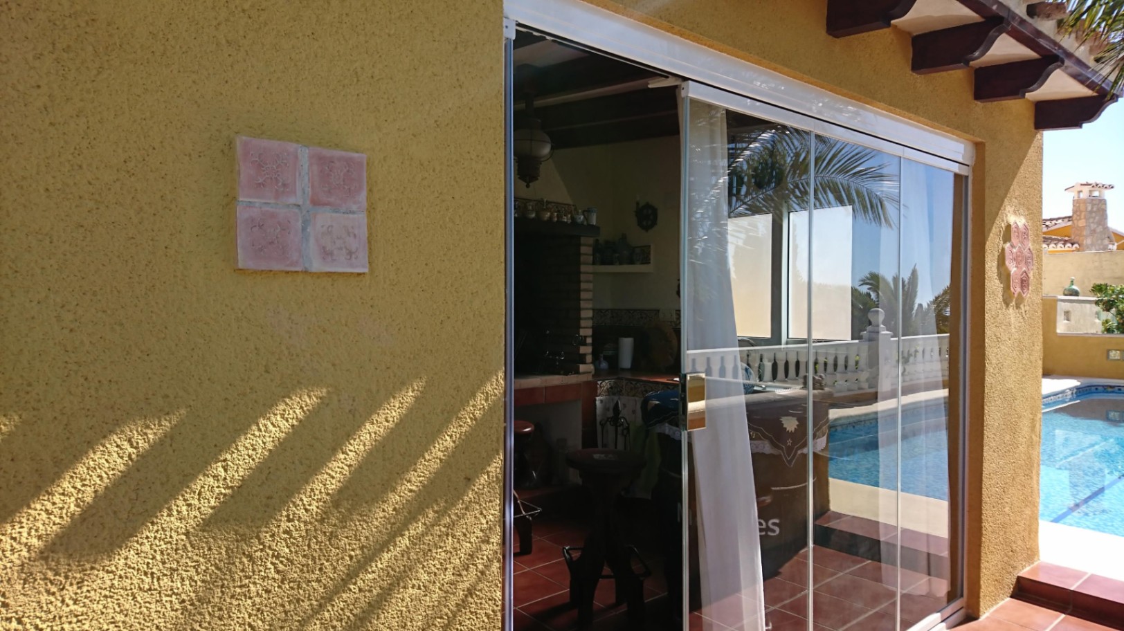 Cocina de verano con barbacoa de obra y vigas de madera, protegida por cerramientos de cristal frente a la piscina y terraza.
