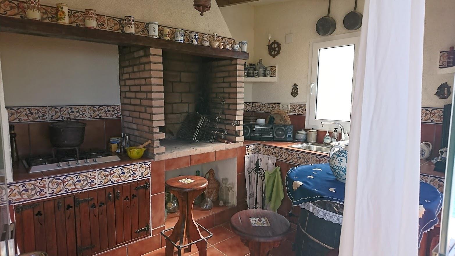 Cocina de verano rústica con barbacoa de obra en ladrillo, encimera de azulejos tradicionales, armarios de madera y fregadero junto a la ventana.
