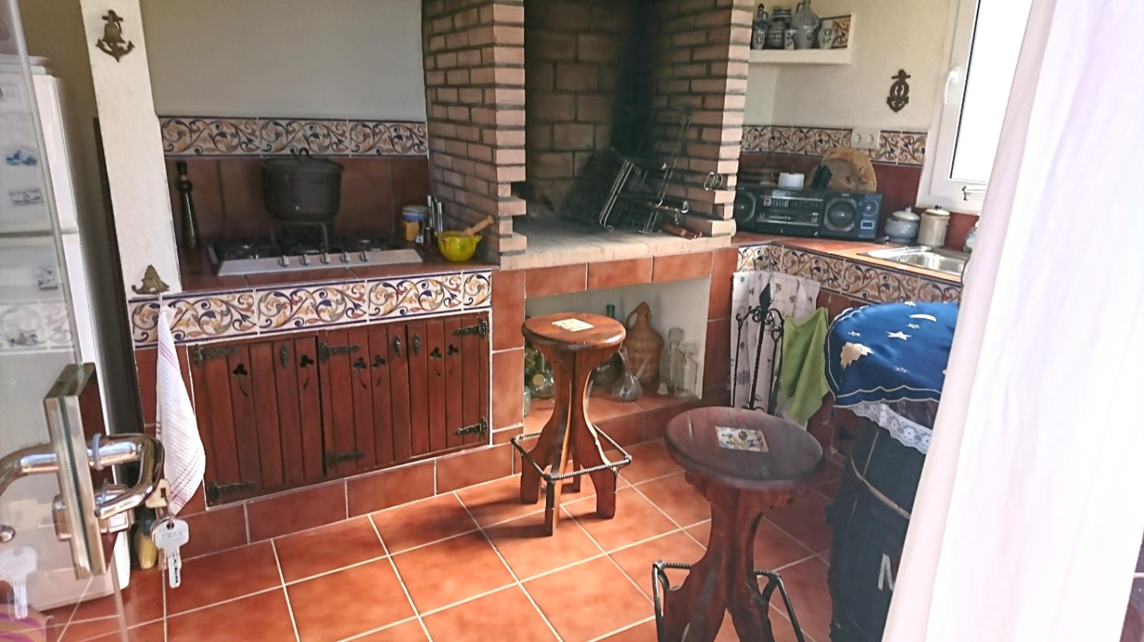 Cocina de verano rústica con barbacoa de obra, encimera de azulejos decorativos, fogones de gas y taburetes de madera sobre suelo de terracota.