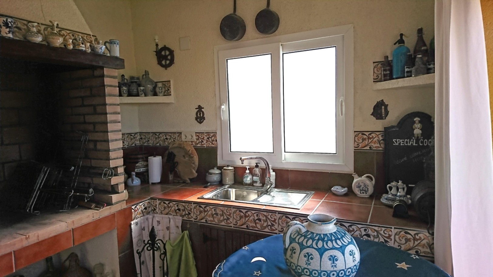 Cocina de verano rústica con barbacoa de obra, encimera de azulejos tradicionales, fregadero de acero inoxidable y ventana de PVC que aporta luz natural.